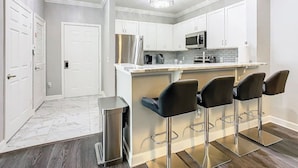 A modern kitchen with white cabinets, a stainless steel refrigerator, a microwave, and a bar area with three black bar stools. - Luxury Modern 3BR Near Disney (Davenport)