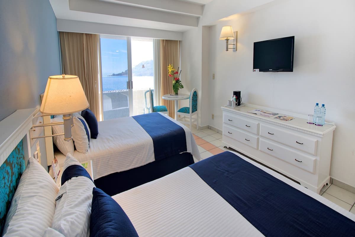 Habitación de hotel con dos camas, televisión, escritorio con lámpara, una silla y vista al mar.
