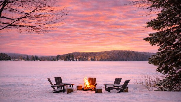 Schneebedeckte Landschaft mit Feuerstelle und zwei Stühlen, die einen Sonnenuntergang über einem zugefrorenen See zeigen.