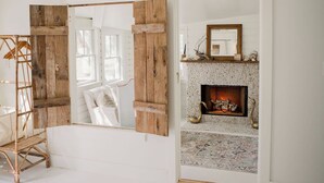 A spacious room with a large bed, a rustic wooden door, a fireplace with decorative items, and a view of the outdoors. - Water Street Cottage (Swansboro)