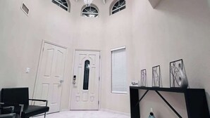 Una habitación amplia con techo alto, un candelabro, una puerta con panel de vidrio y un estante con obras de arte enmarcadas.