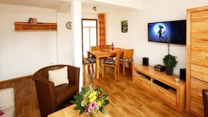 A living room with a brown leather sofa, a wooden coffee table with a floral centerpiece, a dining area with a wooden table and chairs, a flat-screen TV mounted on the wall, and a wooden cabinet with drawers.