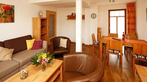 A living room with a brown leather sofa, a wooden coffee table with a vase of flowers, a brown leather armchair, a wooden dining table with chairs, and a wooden bookshelf.