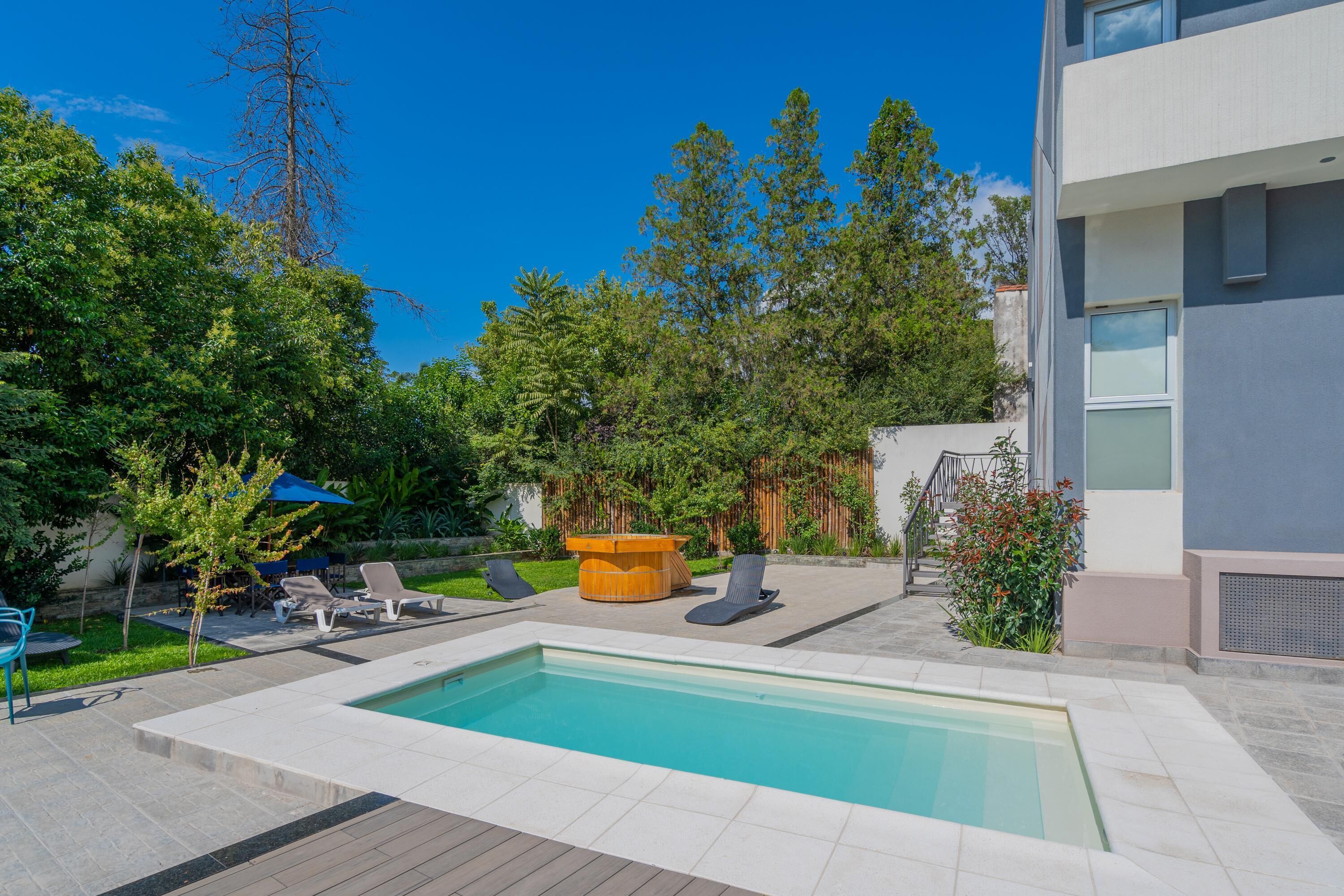 A swimming pool with lounge chairs and a wooden table, surrounded by greenery and a modern building.