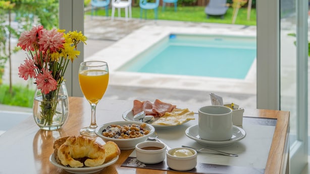 Mesa de desayuno con cruasanes, fruta, café y un vaso de zumo de naranja, con vistas a una piscina y jardín.