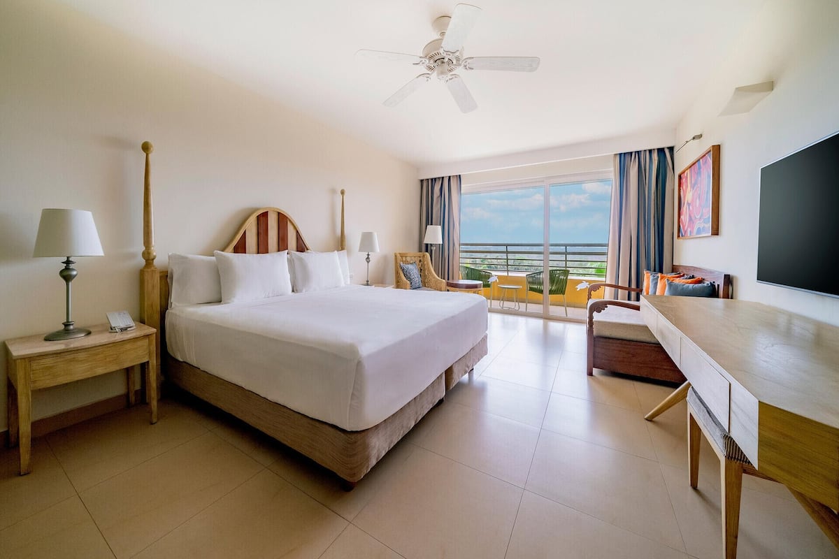 A hotel room with a large bed, a desk, a chair, a nightstand, a lamp, a ceiling fan, and a view of the ocean.