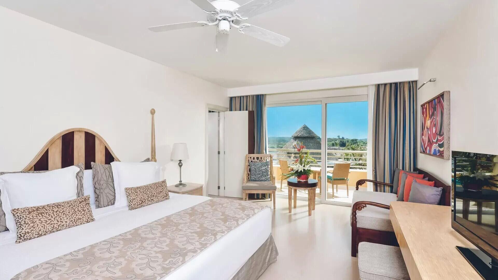 Habitación de hotel con cama, escritorio, silla, televisión y balcón con vistas.
