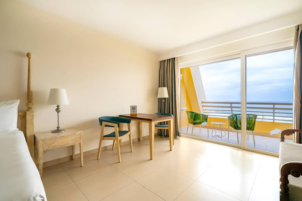 A hotel room with a bed, a desk, two chairs, a lamp, and a balcony with a view of the sea.