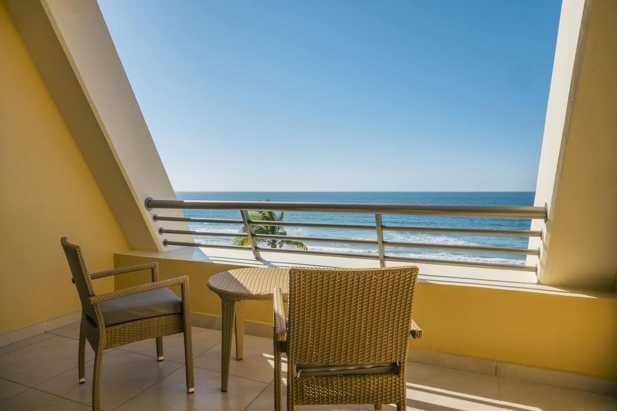 A balcony with two wicker chairs and a small table, overlooking a beach with the ocean and palm trees.