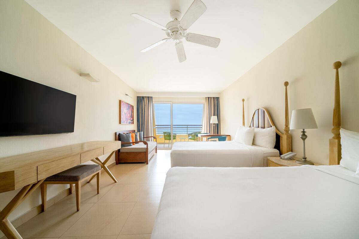 A hotel room with a bed, a desk, a chair, a TV, a ceiling fan, and a view of the ocean.