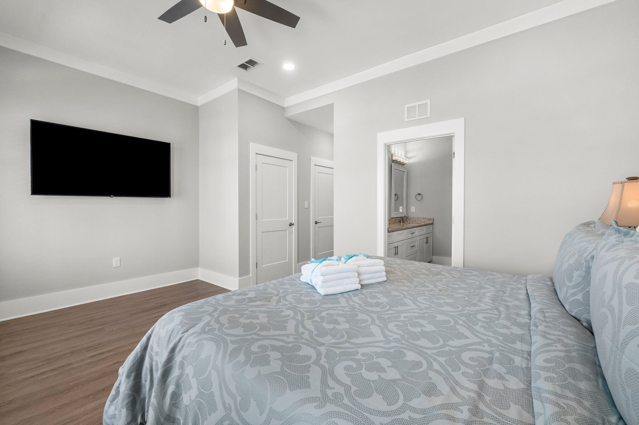 Une chambre dotée d’un grand lit, d’un téléviseur à écran plat fixé au mur, d’un ventilateur de plafond et d’une salle de bain visible à travers une porte ouverte.