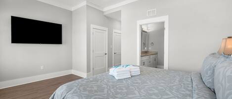Une chambre dotée d’un grand lit, d’un téléviseur à écran plat fixé au mur, d’un ventilateur de plafond et d’une salle de bain visible à travers une porte ouverte.