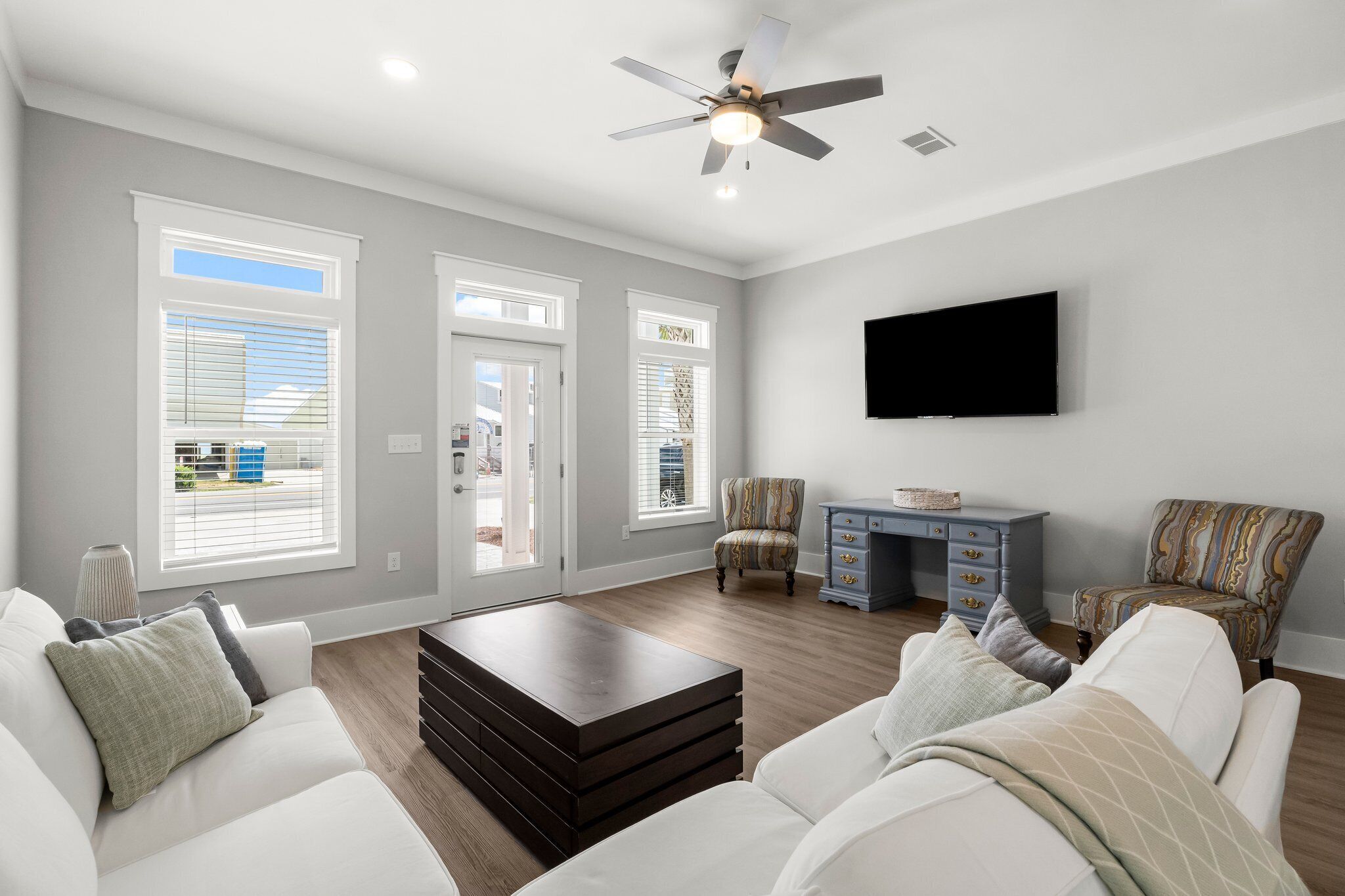 A spacious living room with a large window, a ceiling fan, a flat-screen TV, and a desk with a chair.