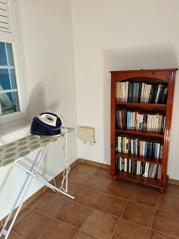 A room with a wooden bookshelf filled with books, a window with blinds, and an ironing board with an iron.