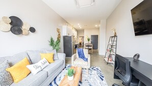A modern living room with a grey sofa, a wooden coffee table, and a black office chair. There is a TV mounted on the wall and a small shelf with decorative items. - King Bed, Free Parking, Near Stadium (Cincinnati)