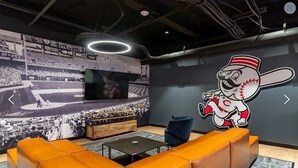 A modern lounge area with a large mural of a baseball stadium, a television, and a mascot illustration on the wall. - King Bed, Free Parking, Near Stadium (Cincinnati)