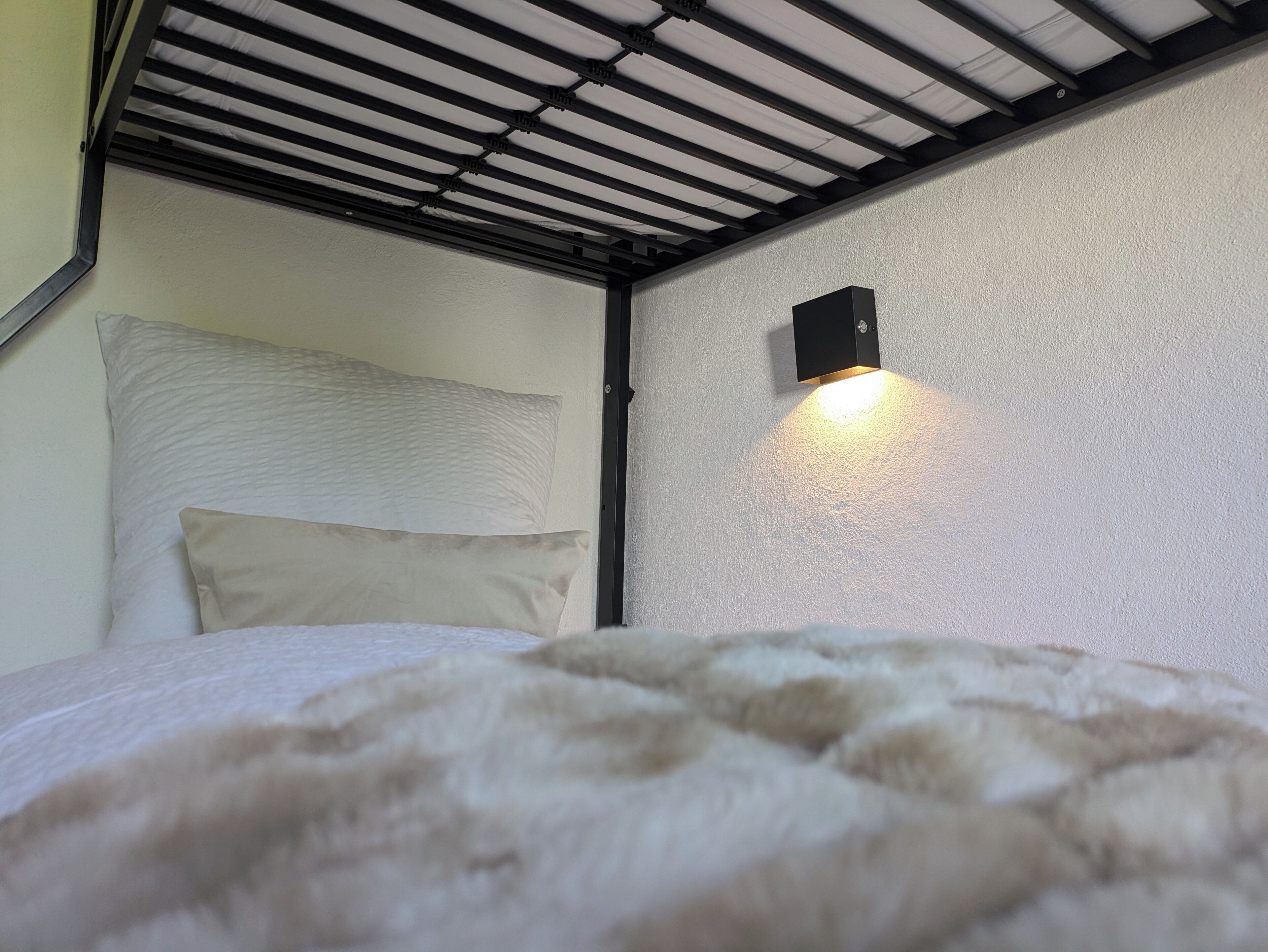 Uma cama beliche com cabeceira e almofadas, teto com grade de metal e luminária fixada na parede.