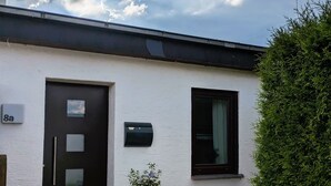 En vit byggnad med en svart dörr och ett svart fönster, en brevlåda och en krukväxt.