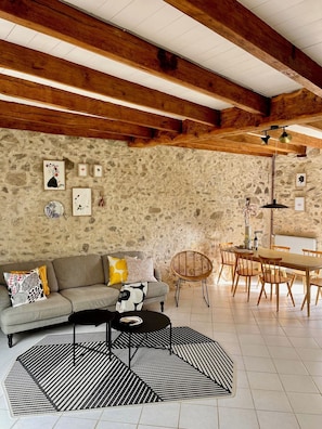 Un salón acogedor con pared de piedra, vigas de madera, un sofá, una mesa de comedor y una alfombra estampada.