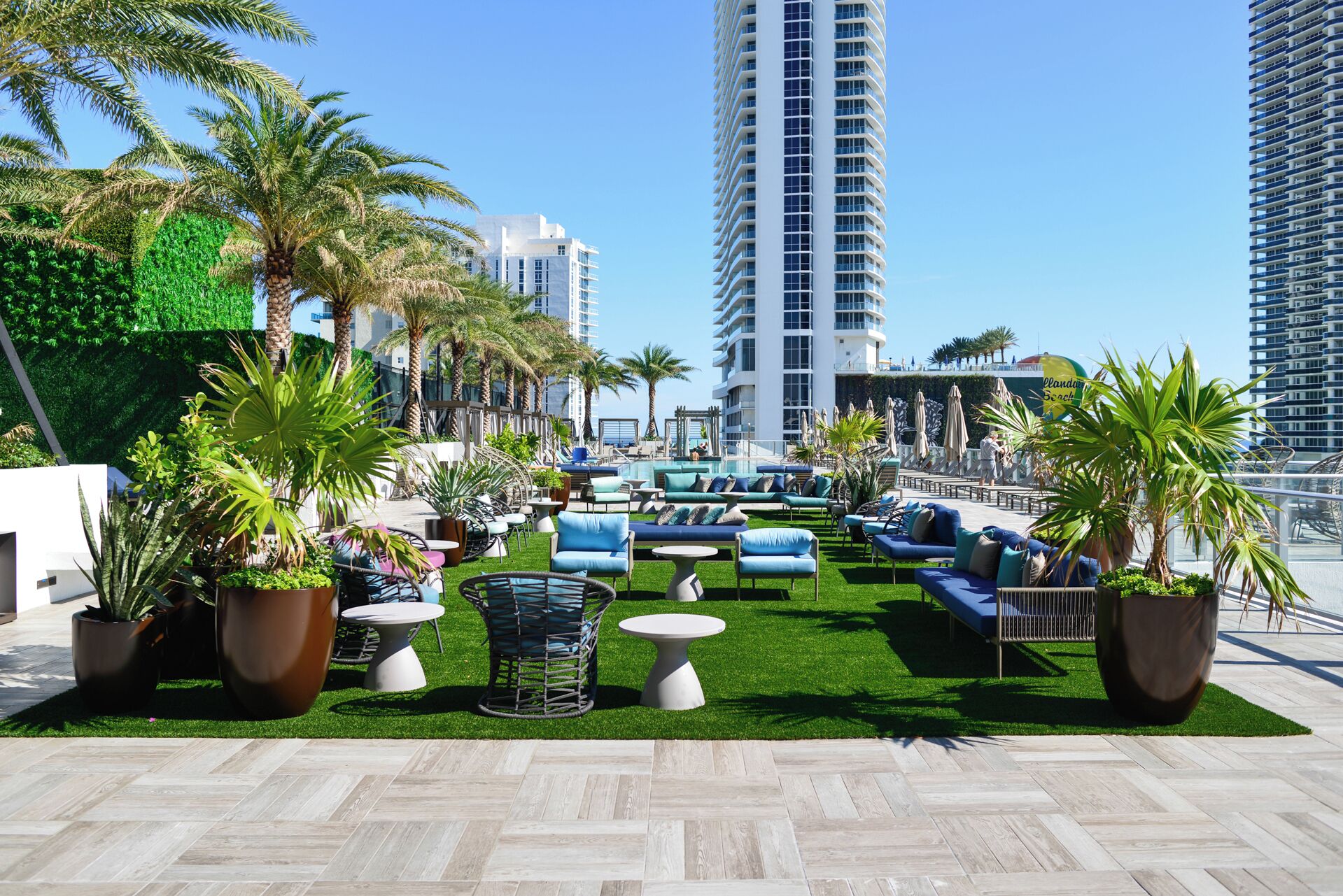 A rooftop lounge with outdoor seating, palm trees, and a view of high-rise buildings.
