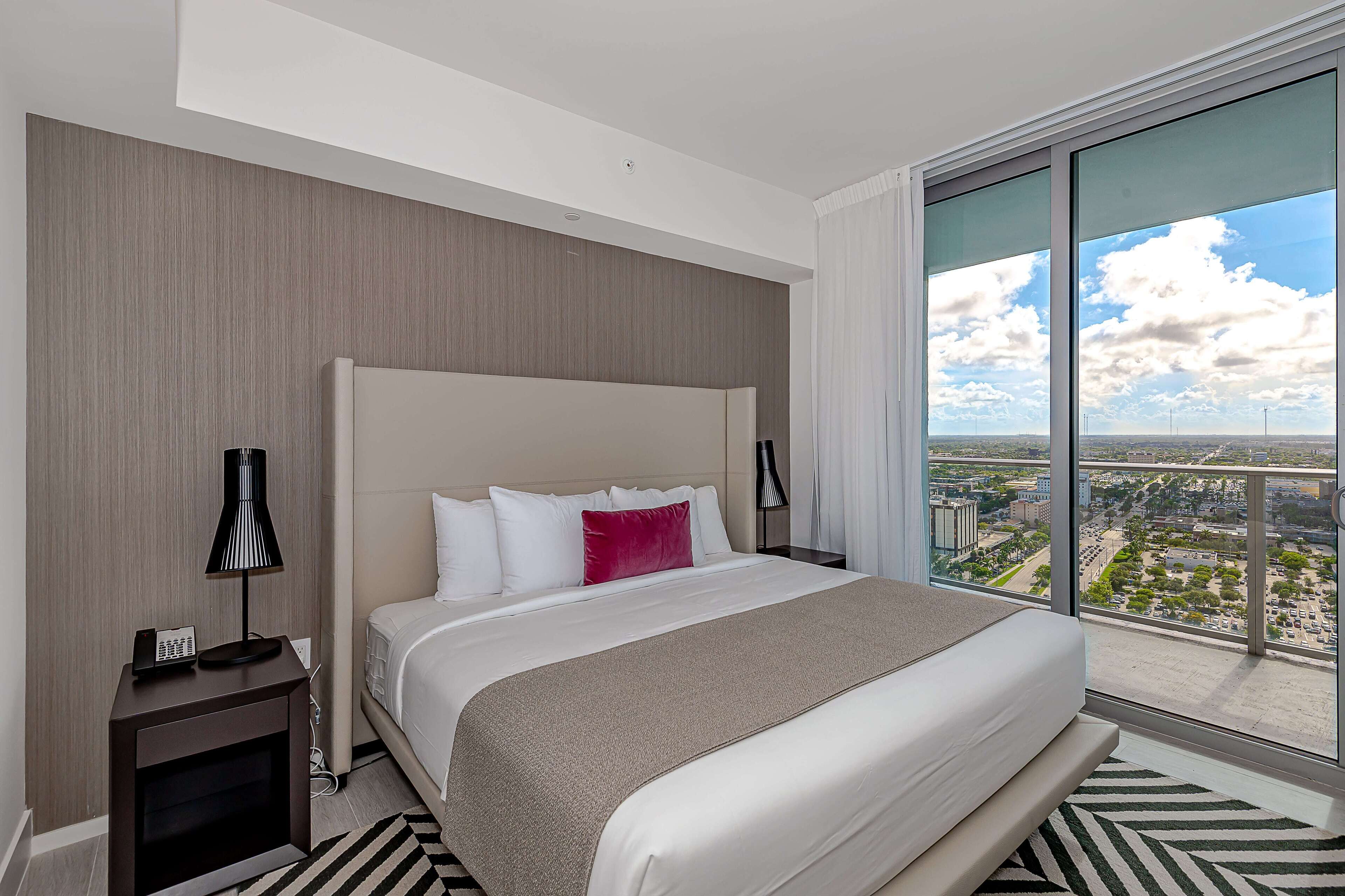 A modern hotel room with a large bed, a nightstand, a lamp, and a view of the city through large windows.