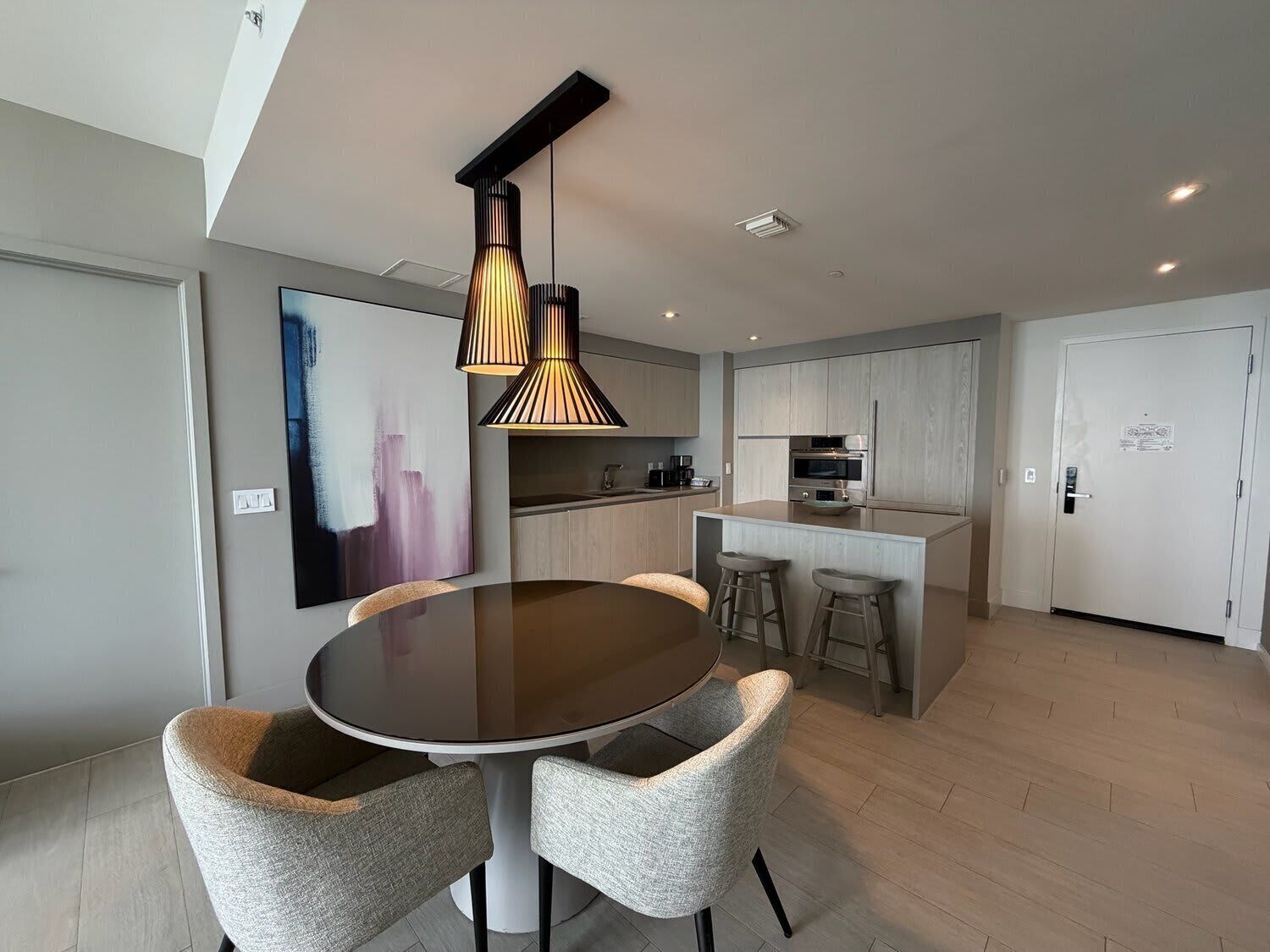 A modern kitchen with a dining area, featuring a round table with four chairs, pendant lighting, and a large abstract painting on the wall.