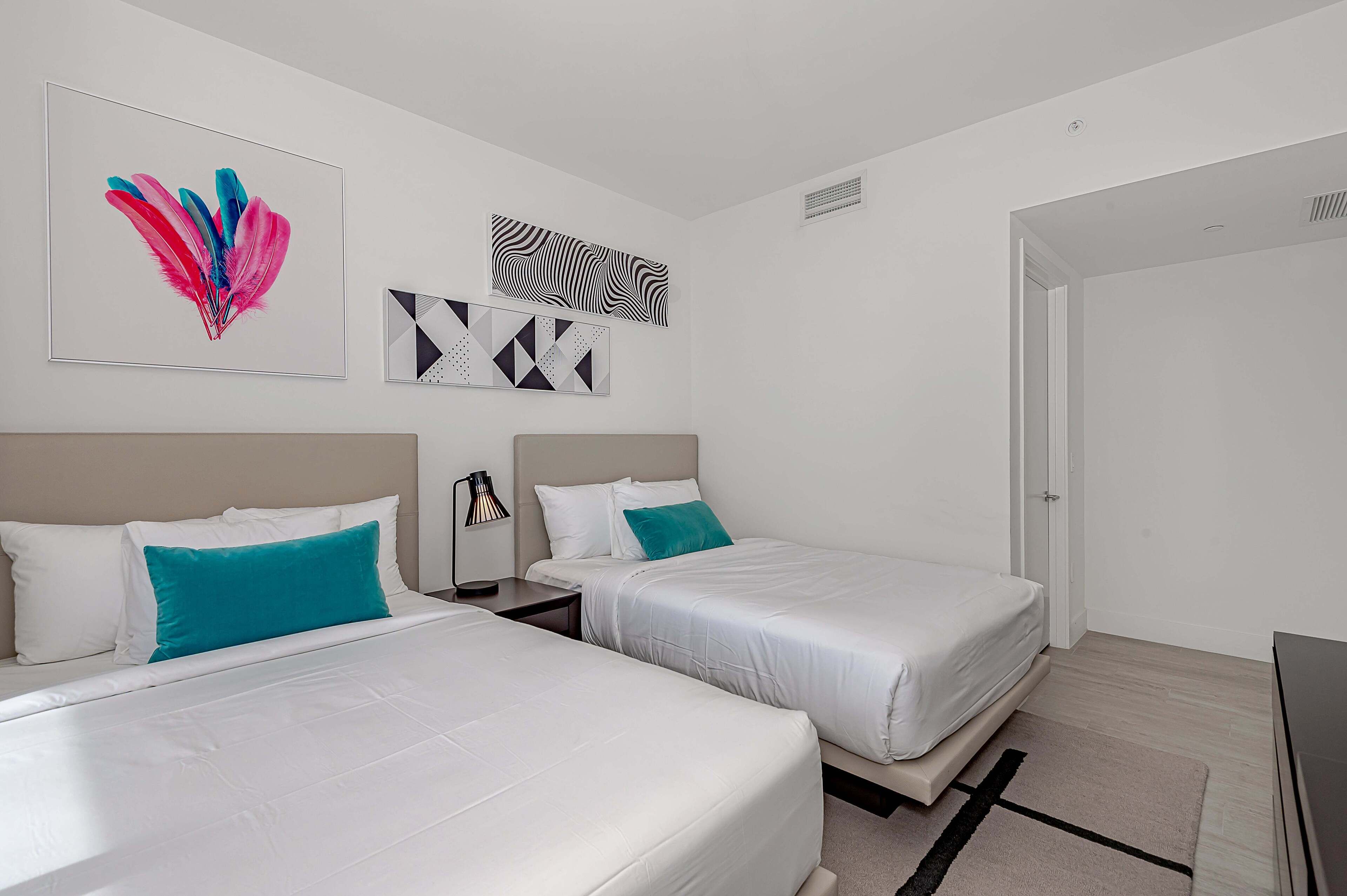 A hotel room with two beds, white bedding, teal pillows, and geometric wall art.