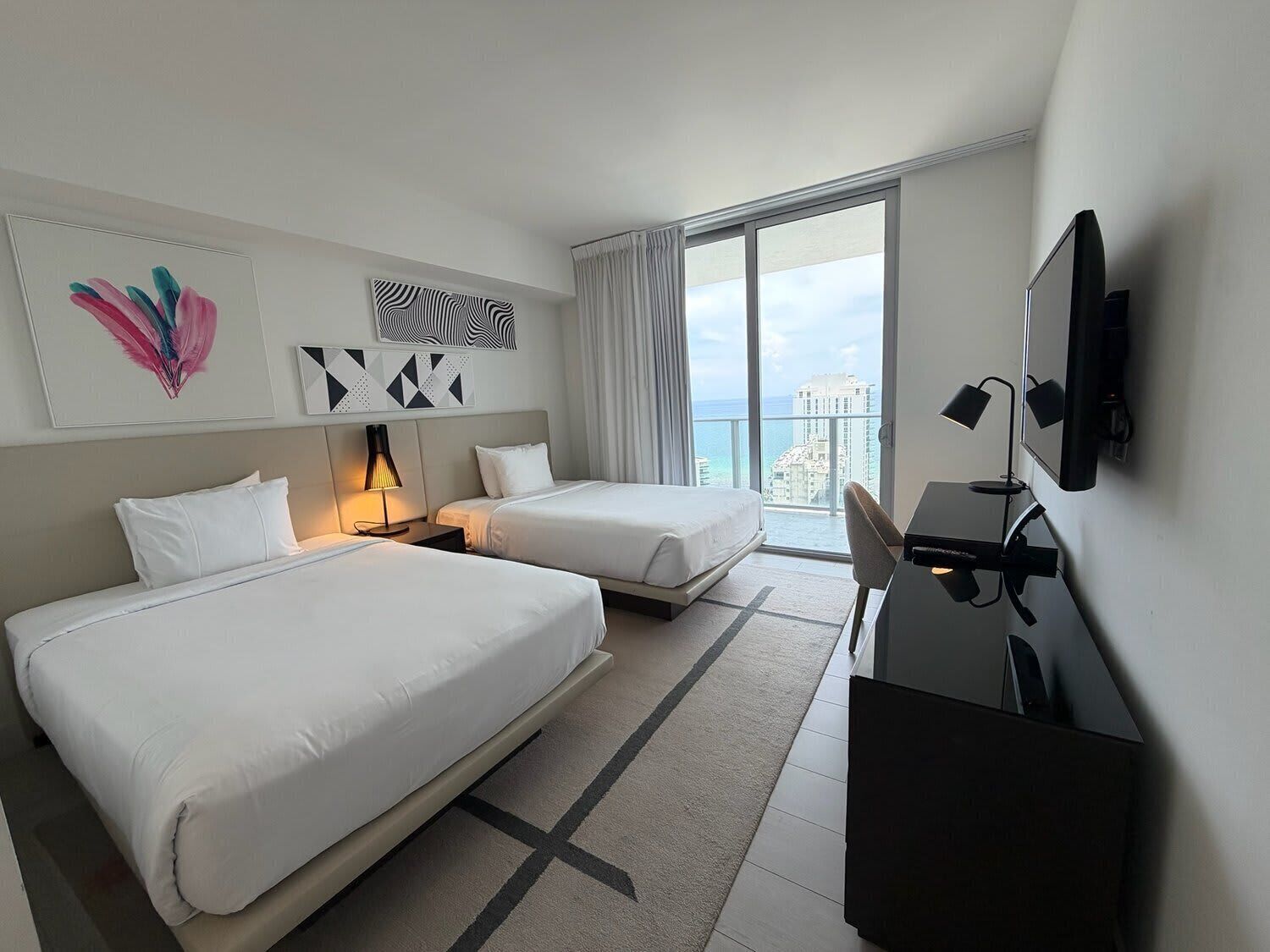 A hotel room with two beds, a desk with a TV, a large window with a city view, and artwork on the walls.
