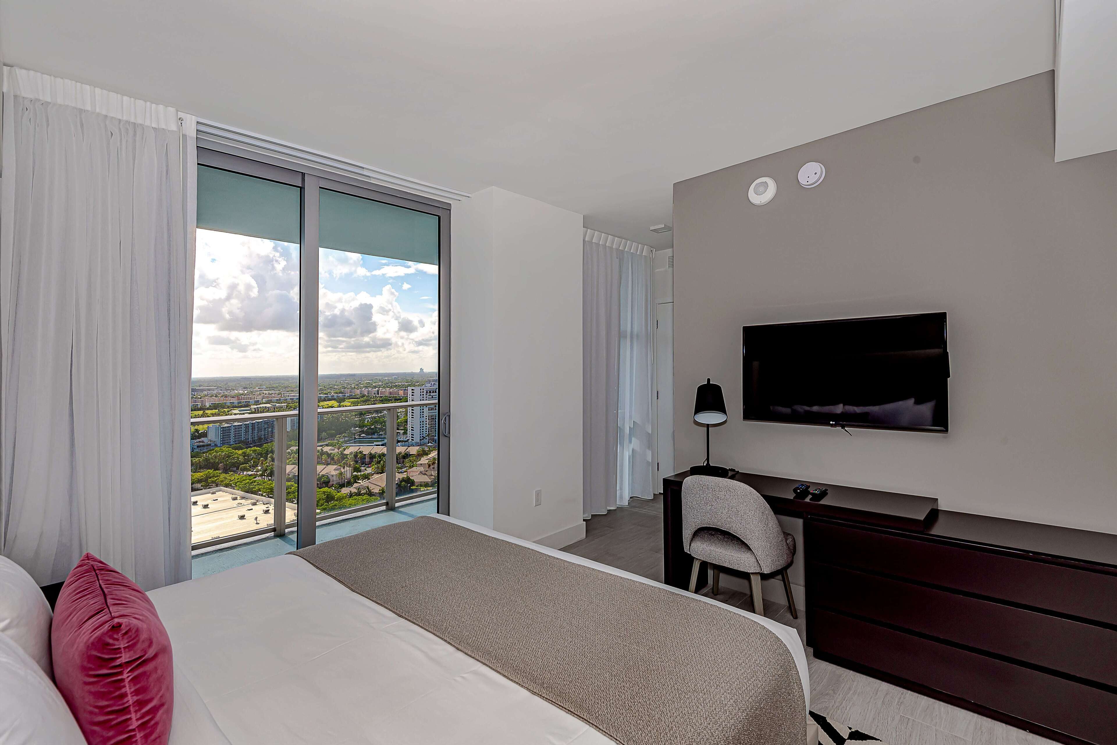 Habitación de hotel con una cama grande, un televisor montado en la pared, un escritorio con una silla y vistas de la ciudad a través de amplias ventanas.
