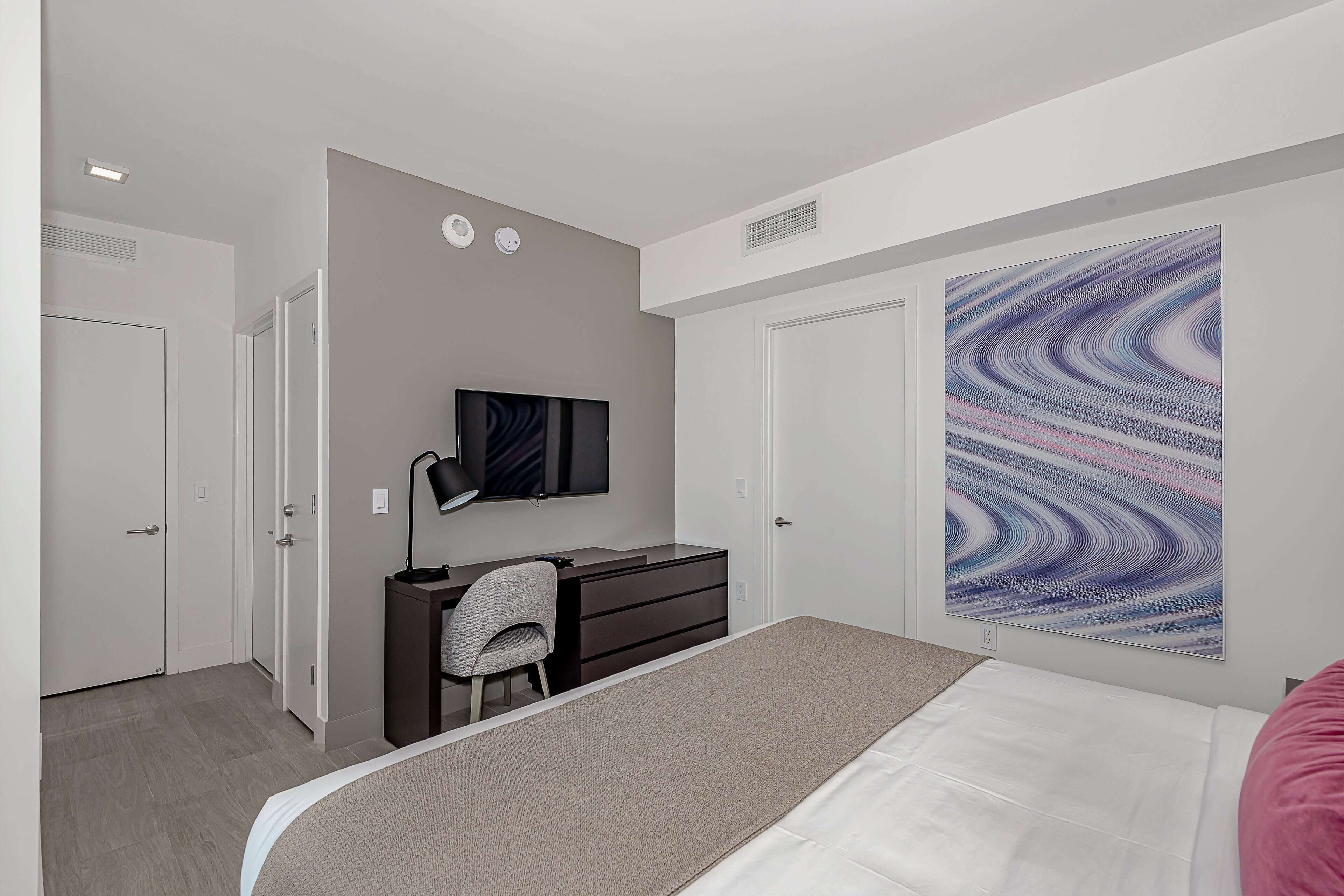 A bedroom with a bed, a desk with a chair, a television, and a large abstract painting on the wall.