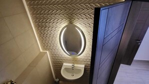 Un baño moderno con techo de diseño en patrón espiga, un espejo redondo, lavabo pedestal y un inodoro.