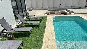 Una piscina con agua azul cristalina, rodeada por una plataforma de concreto y una pequeña zona con césped. Hay sillas de descanso y una cesta sobre el césped.