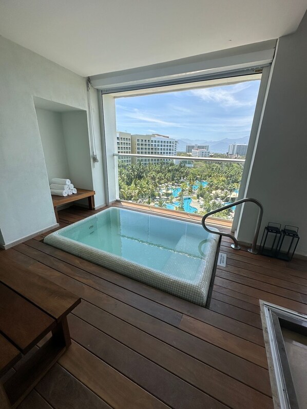 Un balcon doté d’un jacuzzi à débordement protégé par des parois en verre, d’un plancher en bois et offrant une vue sur un complexe hôtelier comprenant plusieurs piscines et bâtiments.