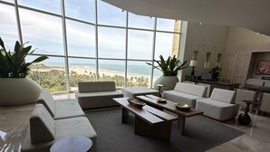 A spacious living room with a large window overlooking a beach, featuring a sofa, coffee table, and a dining area.
