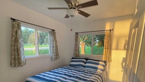 Un dormitorio con una cama, dos ventanas con cortinas, un ventilador de techo y una puerta.