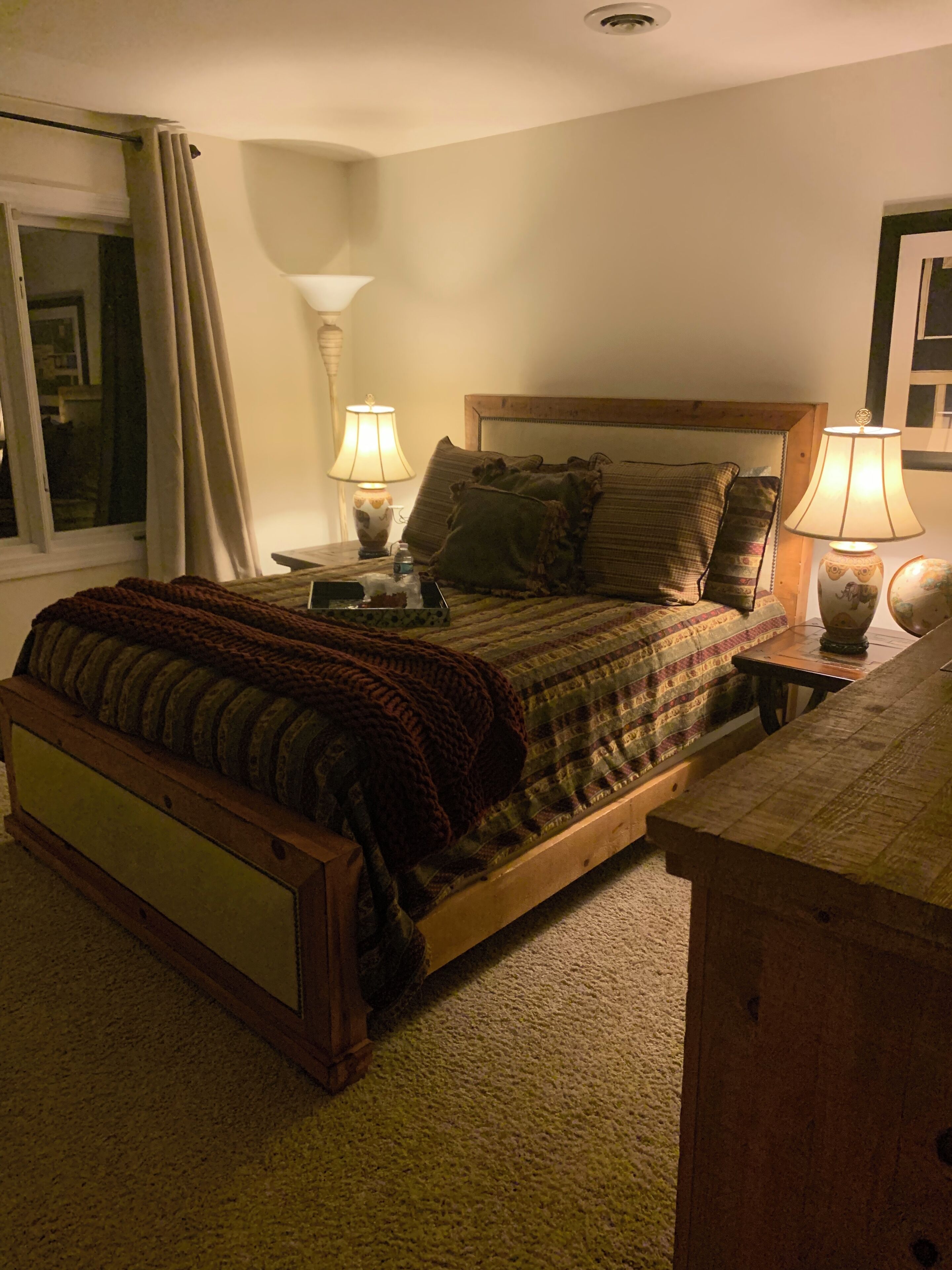A bedroom with a wooden bed, two bedside lamps, a nightstand, a window with curtains, and a view of the outside.