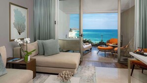 A modern hotel room with a balcony overlooking the ocean, a sofa, a small table, and a view of the beach. - Grand Velas Los Cabos - All Inclusive (Cabo San Lucas)
