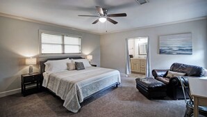 Ein Schlafzimmer mit einem Bett, Nachttischen, einem Deckenventilator, einem Fenster mit Jalousien, einem Gemälde, einem Ledersessel und einem Schreibtisch.