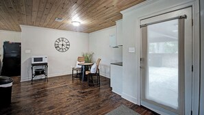 Ein Zimmer mit Holzboden, einer Wanduhr, einer Mikrowelle, einem kleinen Tisch mit Stühlen und einer Glastür zum Außenbereich.