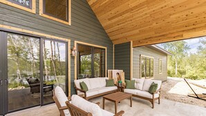 Een overdekt terras met houten plafond, grote schuifdeuren van glas en een witte bank met groene kussens.