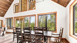 Een eetruimte met een houten tafel en stoelen, grote ramen met uitzicht op bomen en een houten plafond.
