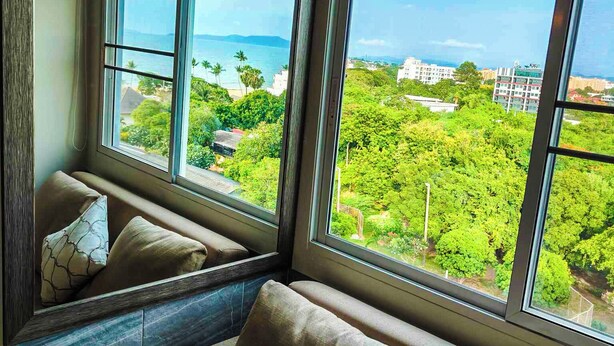 Ein Zimmer mit einem großen Fenster, das einen Blick auf eine grüne Landschaft und Gebäude freigibt. Es gibt eine Couch mit Kissen und eine Klimaanlage über dem Fenster.