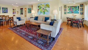 A spacious living room with a sofa, coffee table, and chairs, a dining area with a table and chairs, and various paintings on the walls.