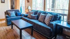 Een gezellige woonkamer met een blauwe zithoek, een houten salontafel en grote ramen met uitzicht op het bos.