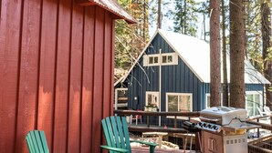 Een houten terras met Adirondack-stoelen, een barbecue en een blauwe blokhut met wit dak.