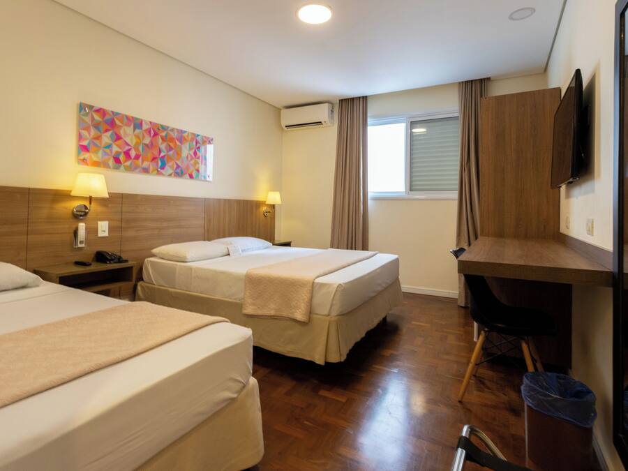 Quarto de hotel com duas camas, uma escrivaninha, uma cadeira, uma televisão e uma janela com cortinas.