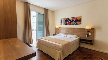 A hotel room with a wooden bed, a desk, a colorful abstract painting, and a view through a glass door.