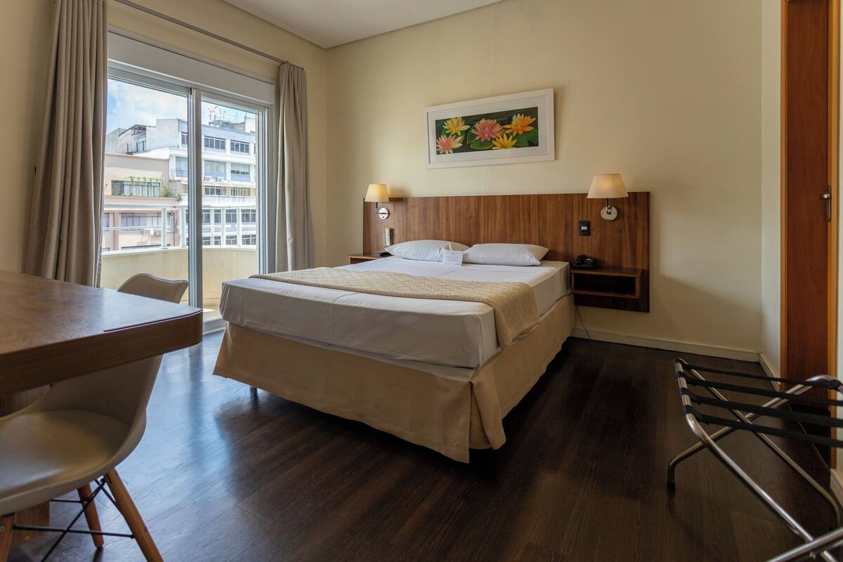 A hotel room with a large bed, a desk, a chair, and a view of buildings through the window.