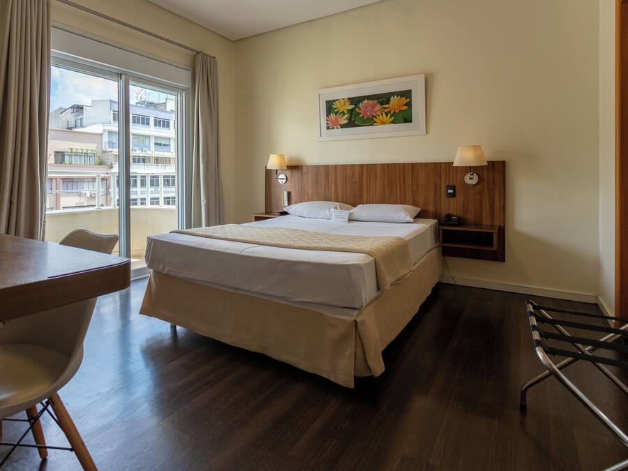 Ein Hotelzimmer mit einem großen Bett, einem Schreibtisch, einem Sessel und Blick auf Gebäude durch das Fenster.