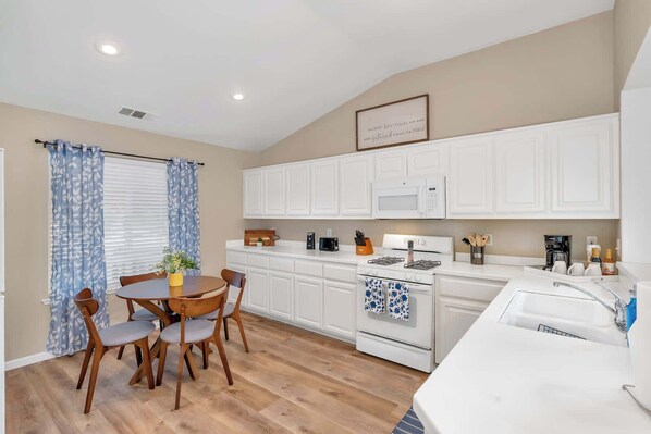 A modern kitchen with white cabinetry, a gas stove, and a dining area with a round table and chairs. - Yosemite Gateway Home Near UC Merced Sleeps 6 (Merced)